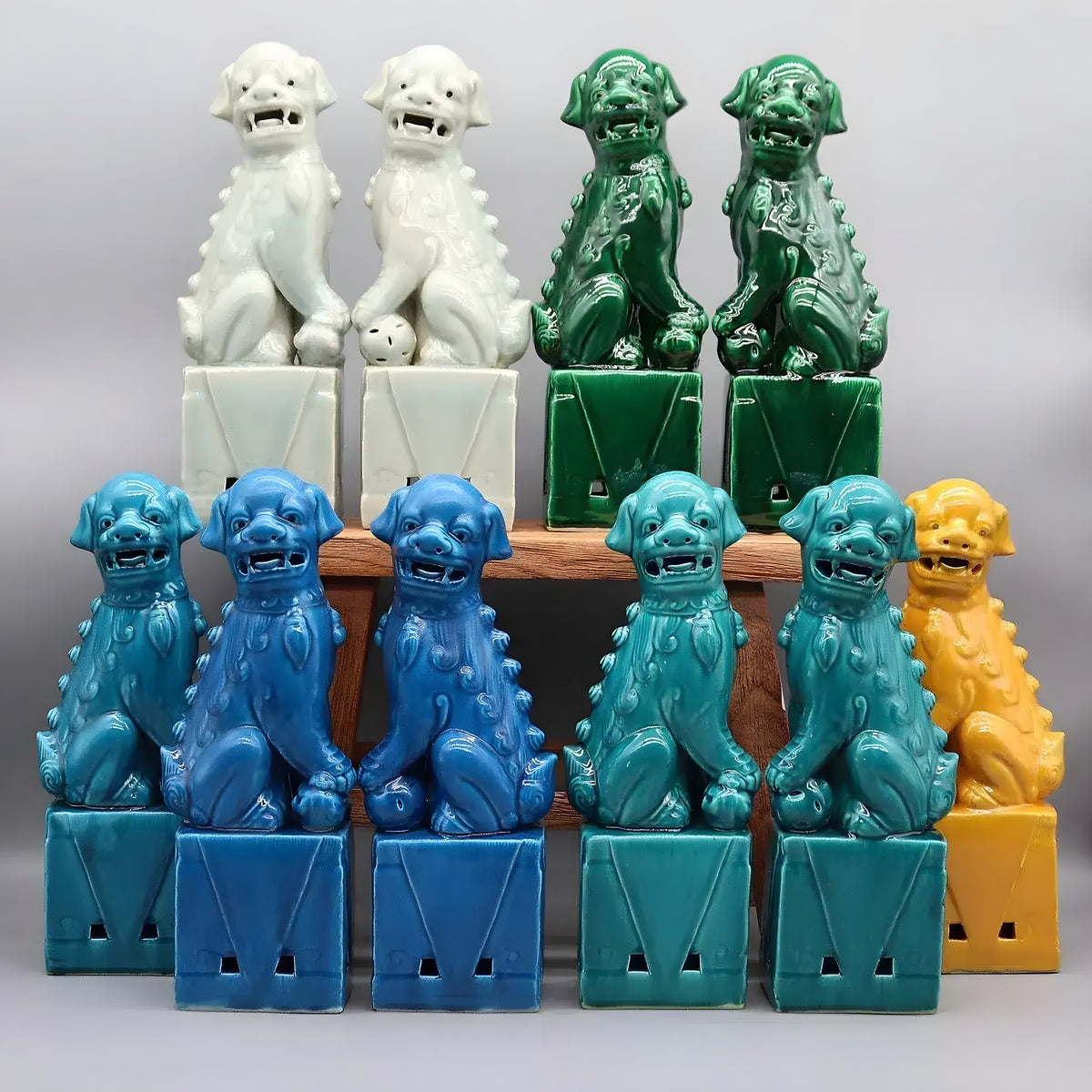 Multi-colorway foo dog ceramic figurine pairs — white, dark green, cerulean, teal, and yellow — on a wood shelf, grey background