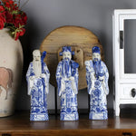 Three blue-and-white Chinese gods hand painted ceramic figurines on dark wood shelf in styled room with terracotta vase and red flowers