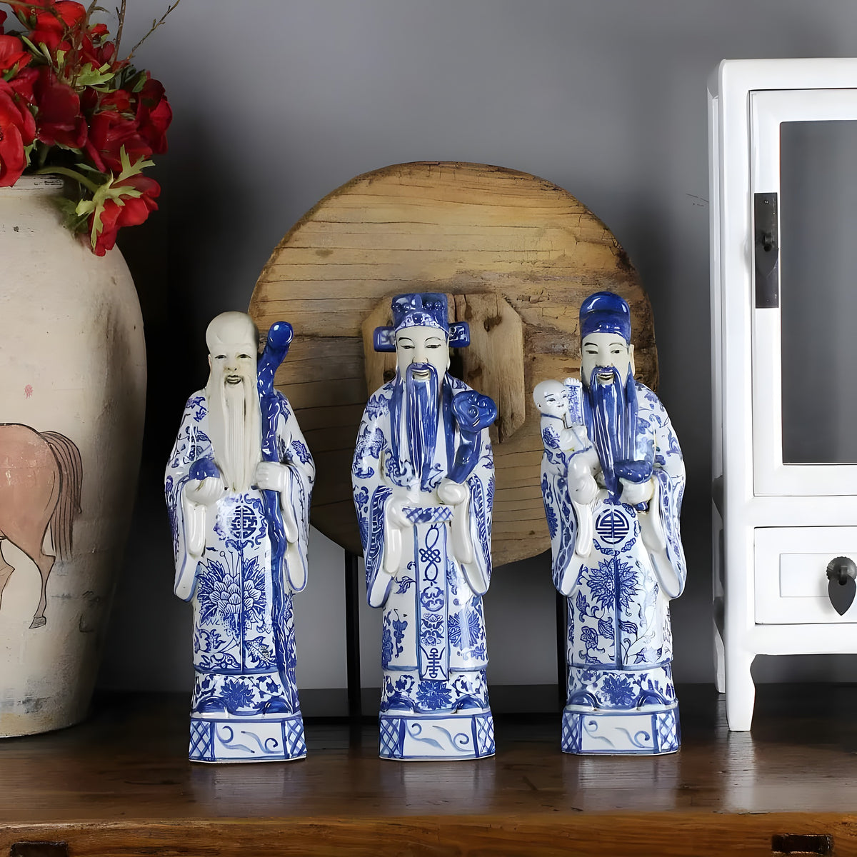Three blue-and-white Chinese gods hand painted ceramic figurines on dark wood shelf in styled room with terracotta vase and red flowers