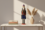 A decorative nutcracker figurine stands on a white marble-top table against a warm beige wall with strong sunlight and shadows. A stack of three books sits on the left, and a beige vase with dried pampas grass is on the right. The nutcracker is dressed in a red and blue uniform with a gold crown and black cape.