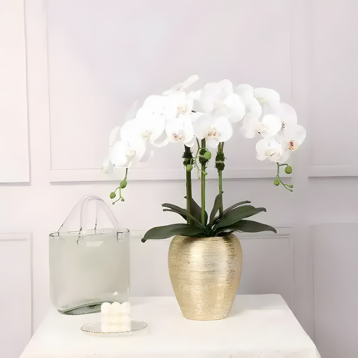 Artificial orchid plant with white phalaenopsis blooms in a textured gold oval pot, clear glass vase alongside, white paneled wall