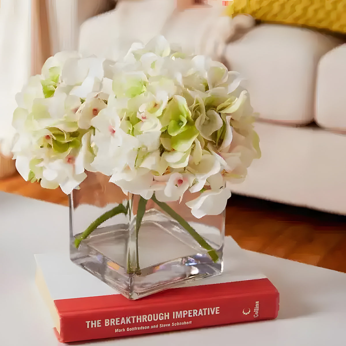 Faux hydrangea centerpiece in square glass vase, white-green blooms on coffee table with book