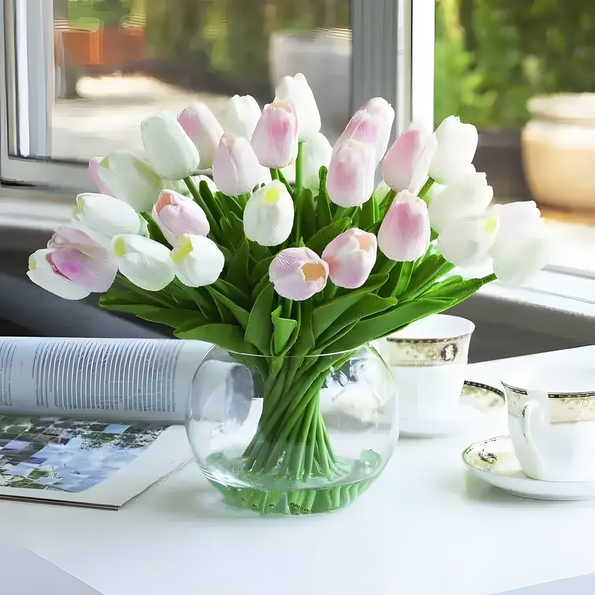 Artificial tulip arrangement in round globe glass vase, mixed white and blush pink tulips, styled on white table with open book