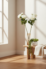 A styled interior scene with white flowers in a tall clear vase and a small potted trailing succulent on a light wood bench, set against a warm beige wall with sunlight and window shadows.