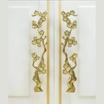 Pair of gold cherry blossom branch door handles on adjacent white cabinet panels, front-facing view
