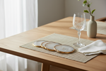A styled dining table setting with woven beige placemats, round gold-edged coasters, a clear wine glass, a folded white napkin, and a small beige vase with greenery on a light wood table.