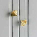 Pair of ginkgo leaf brass door knobs installed on grey-blue painted cabinet doors — polished gold fan-shaped ribbed design