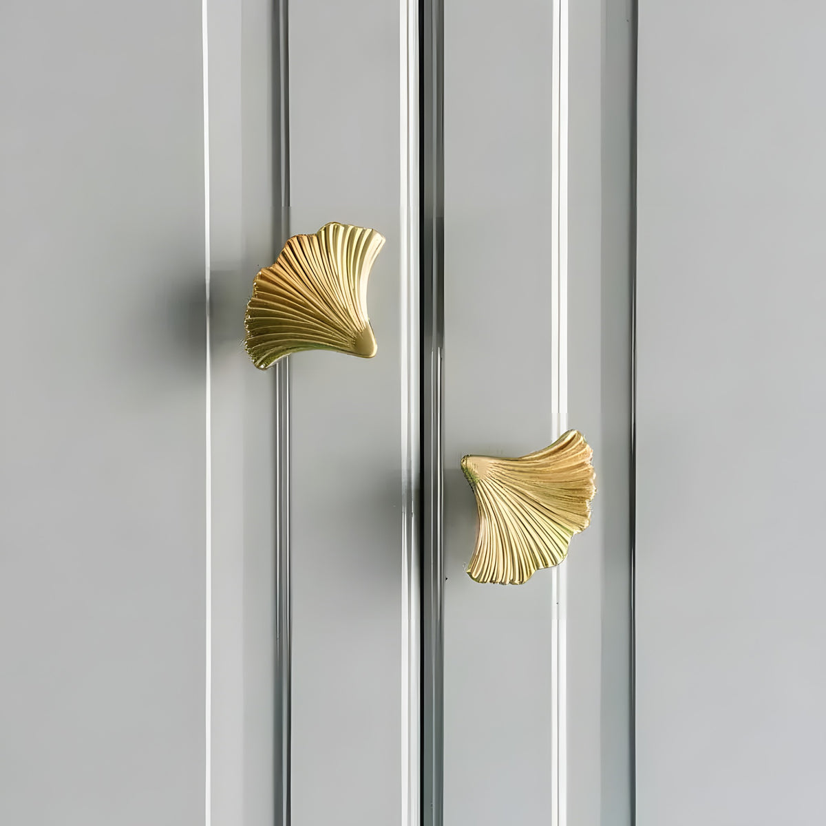 Pair of ginkgo leaf brass door knobs installed on grey-blue painted cabinet doors — polished gold fan-shaped ribbed design