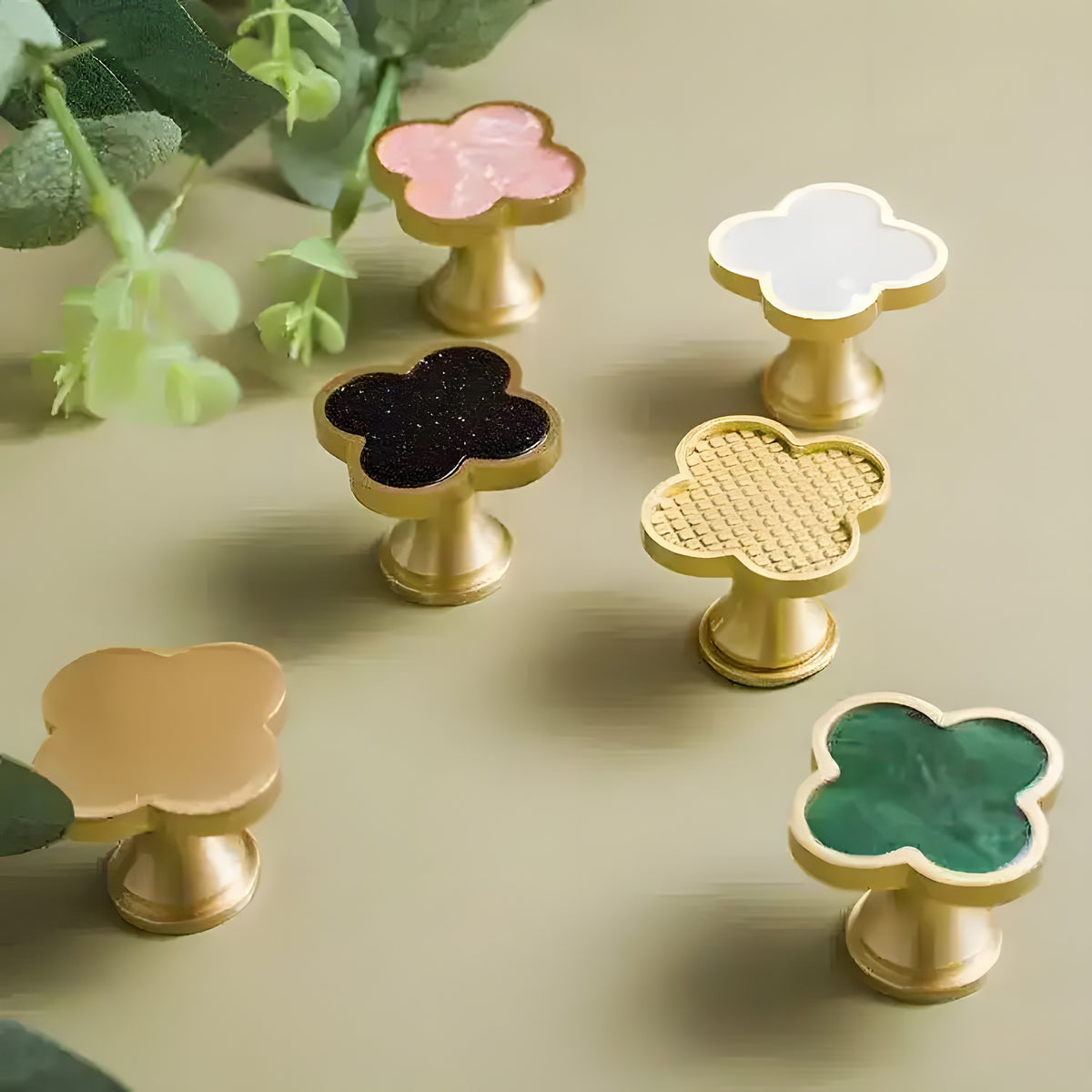 Six brass clover shaped door knobs in pink black white woven nude and green inlay upright on olive green surface