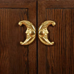 Two brass crescent moon-face drawer knobs mounted facing each other on dark oak cabinet doors