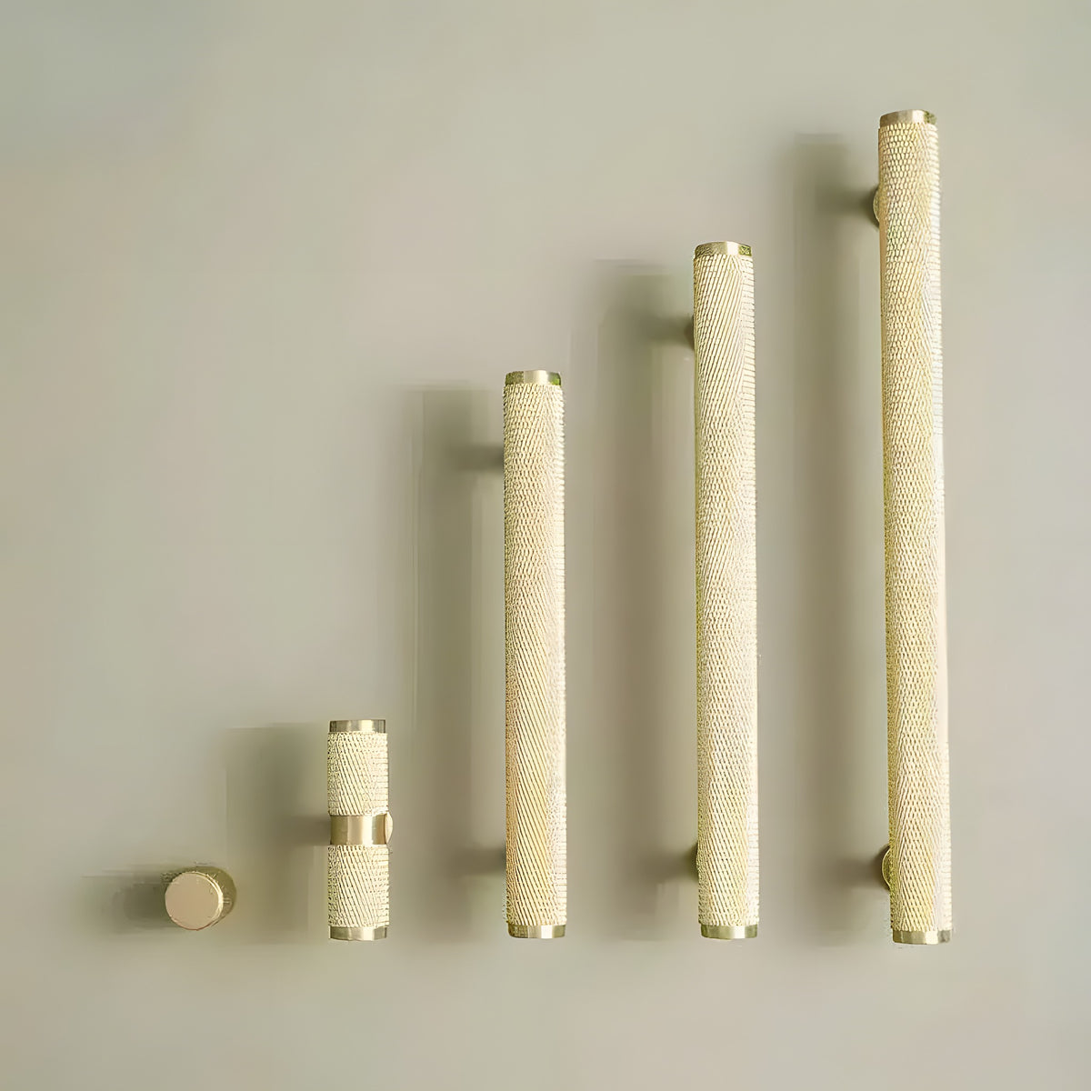 Five textured knurled brass cabinet pulls in graduated sizes from round knob to long bar pull on beige background