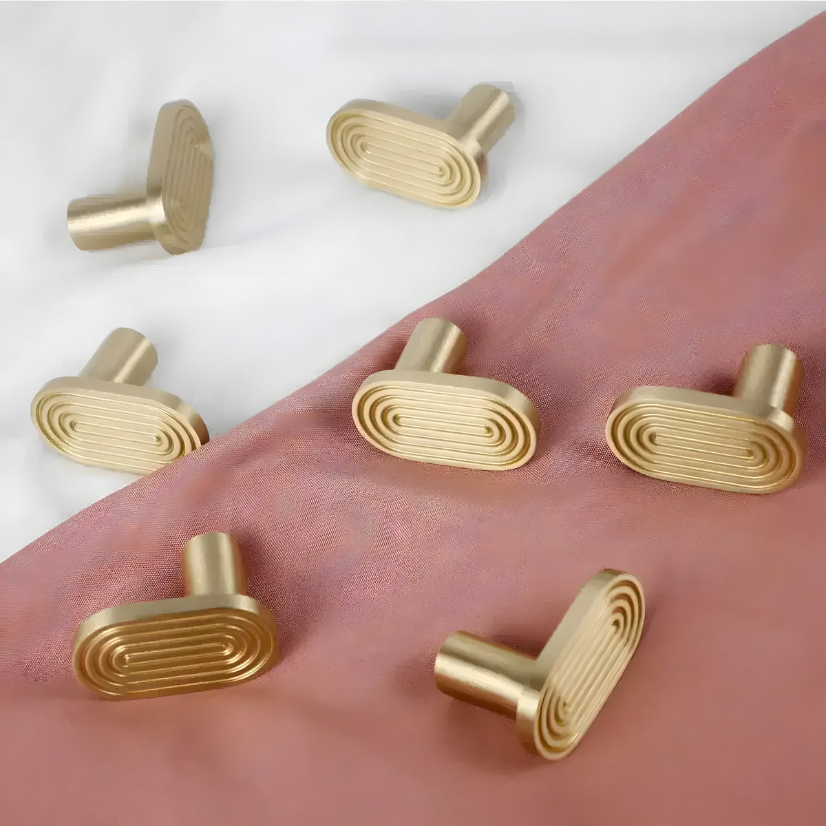 Six matte gold oval art-deco wall hooks scattered on white marble and dusty rose fabric