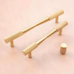 Two brass textured bow-shaped drawer pulls and one cylindrical knob on pink background, knurled barrel grip front view
