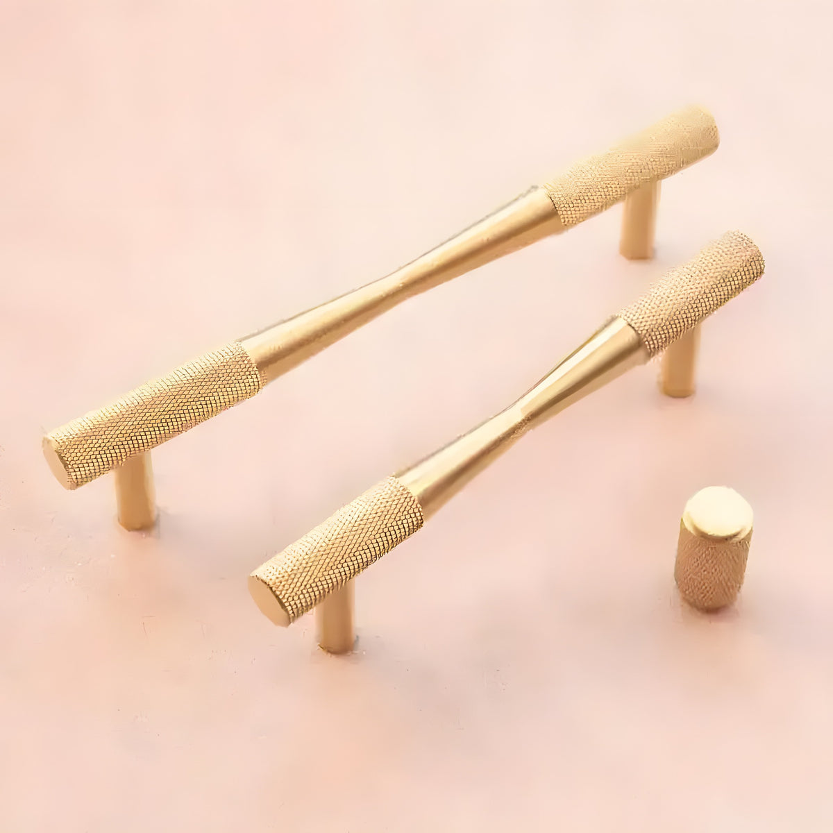 Two brass textured bow-shaped drawer pulls and one cylindrical knob on pink background, knurled barrel grip front view