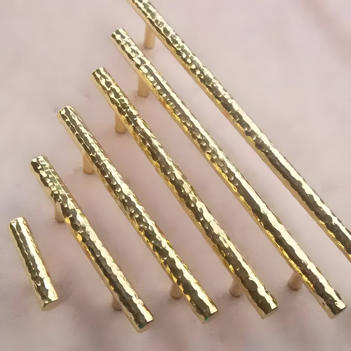 Five hammered brass cabinet drawer pulls in graduated sizes arranged diagonally on pink surface