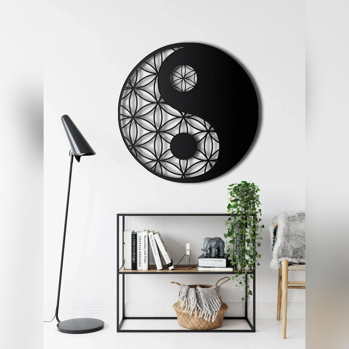 Black round yin yang metal wall art with flower-of-life cutout above open shelf unit, floor lamp and plants alongside