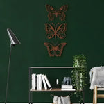Bronze set of 3 butterflies with cutout wings stacked vertically on dark green wall above wire shelf with books and plant