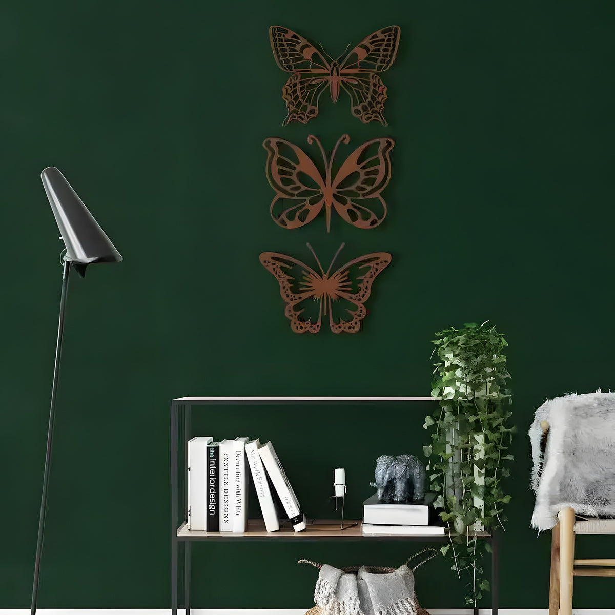 Bronze set of 3 butterflies with cutout wings stacked vertically on dark green wall above wire shelf with books and plant
