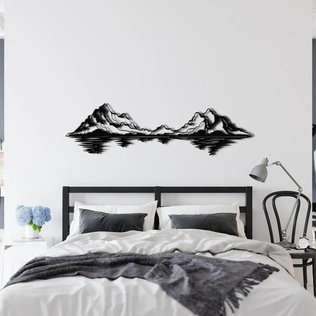Black mountain range landscape metal wall art with lake reflection above dark-headed white bed in minimalist bedroom