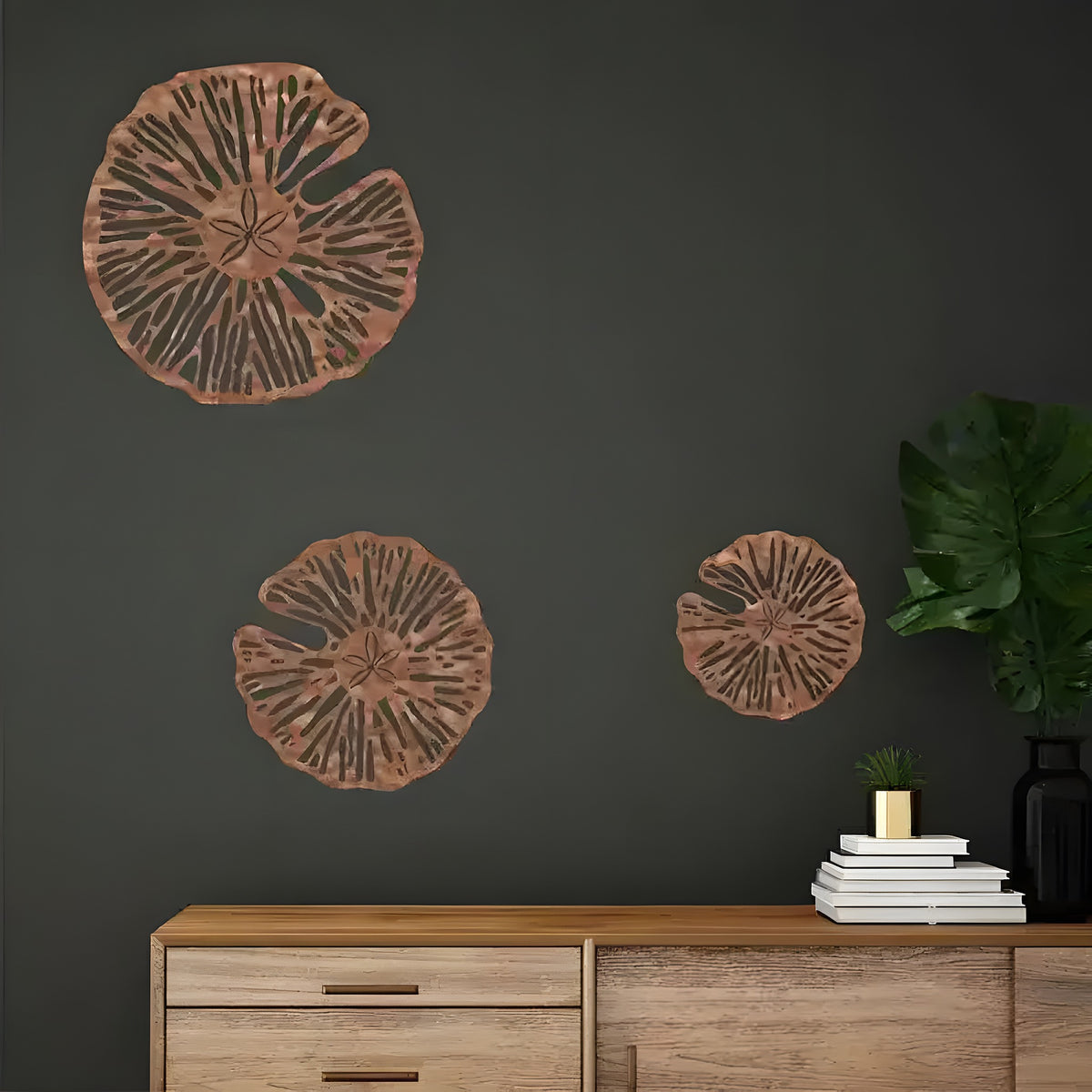 Three copper sand dollar metal wall art pieces in graduated sizes arranged on a dark charcoal wall above oak sideboard