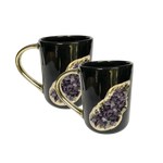 Two black ceramic mugs with gold handles and purple amethyst druzy inset with gold surround, stacked on white