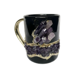 Black ceramic mug with gold handle, large purple amethyst crystal spires above a band of amethyst druzy with gold trim, front view