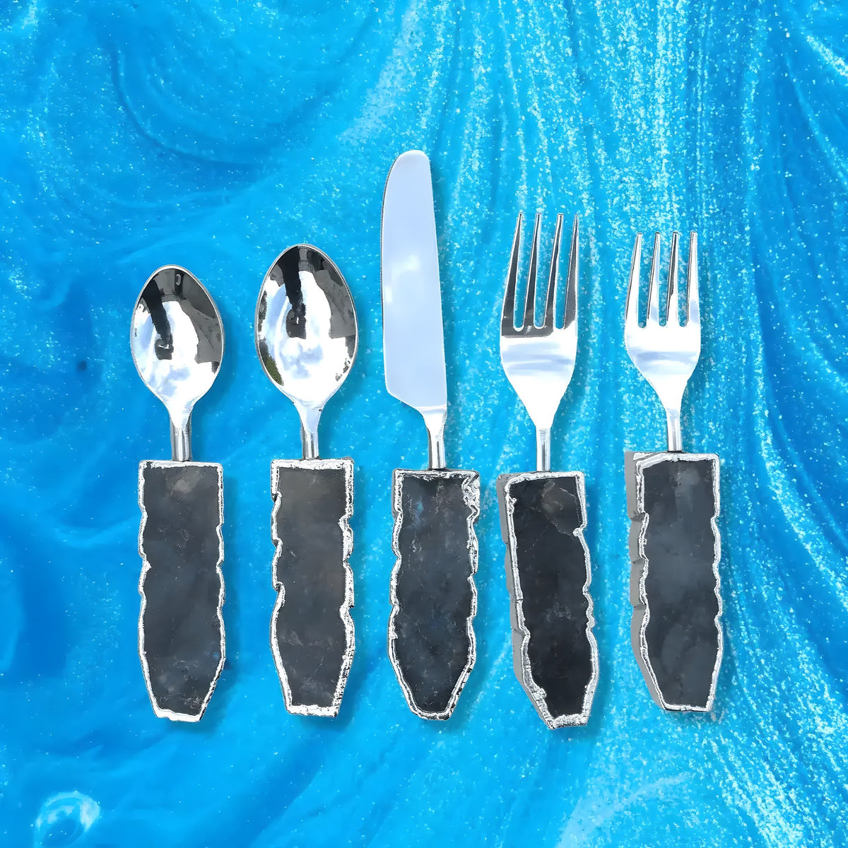 Five-piece labradorite agate cutlery set on blue marble: two spoons, knife, two forks with dark labradorite stone handles, silver trim