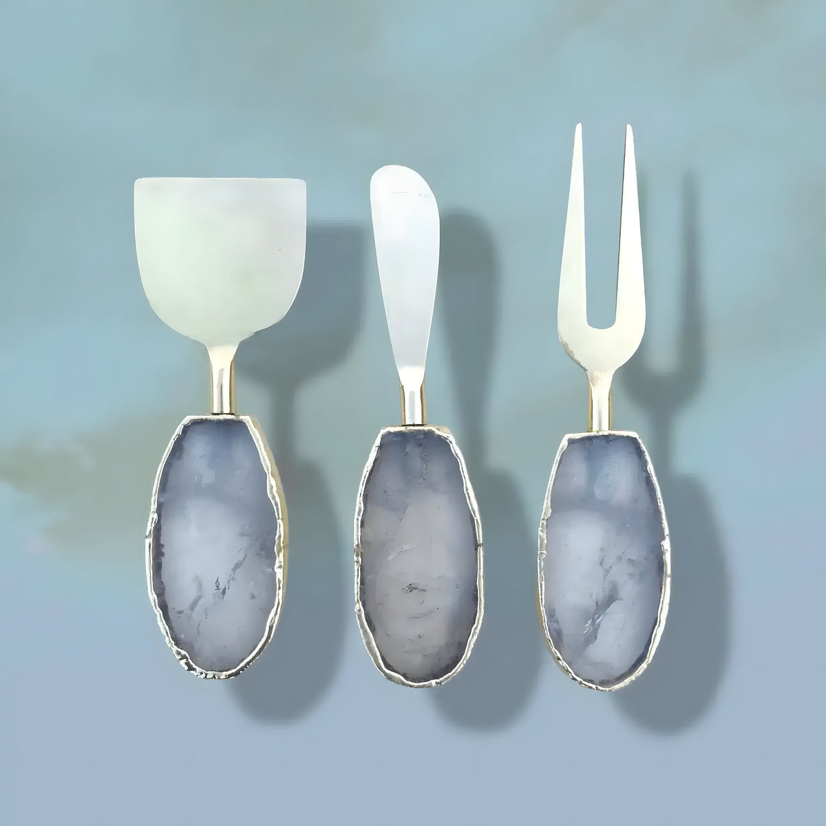 Three silver clear quartz agate cheese tools on blue-grey background, oval grey-clear handles, silver jagged edges