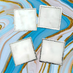 Set of 4 white marble plated coasters, square with silver-edged rims, arranged in two rows on blue and gold marble backdrop