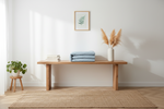 A styled interior scene with a light wood bench against a white wall, a folded stack of pale blue textiles on the bench, a white vase with dried pampas grass, a small potted plant on a stool, and a woven rug on a wood floor.