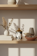 A decorative ceramic jar with a black and white illustrated body and a gold lid sits on a wooden shelf beside a white vase with dried grass, stacked books, a terracotta bowl with stones, and a small gray vase in a softly lit interior setting.