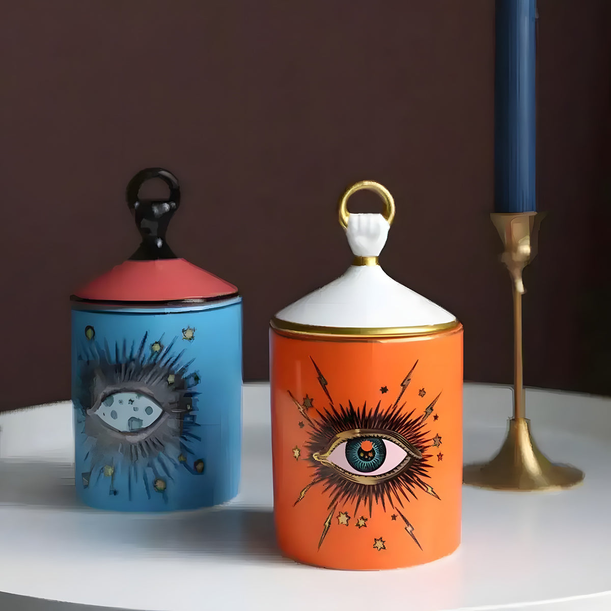 Two magic eye aromatherapy candle jars — blue with black lid and orange with white gold-rimmed lid, cosmic eye motifs