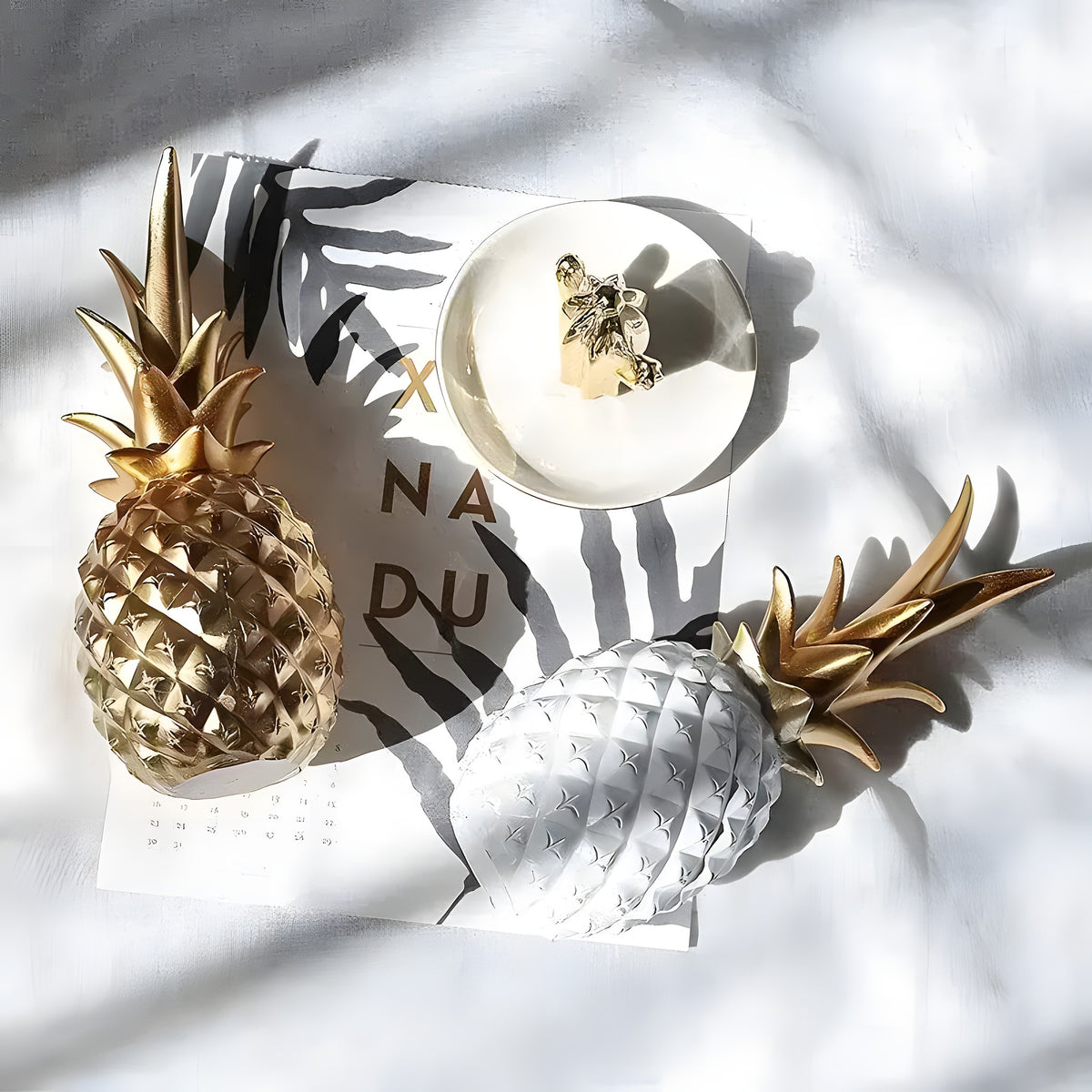 Gold and white tropical pineapple table decor figurines on white fabric flat-lay with candle, casting shadows