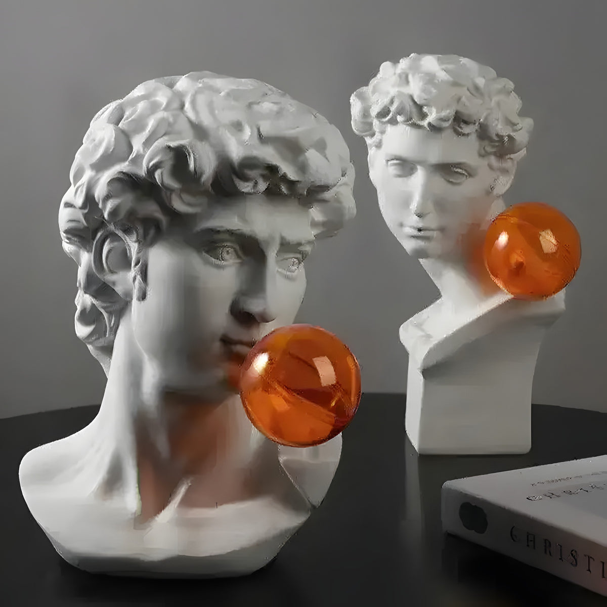 Two white classical Greek god bust sculptures with orange bubble spheres on dark table beside book