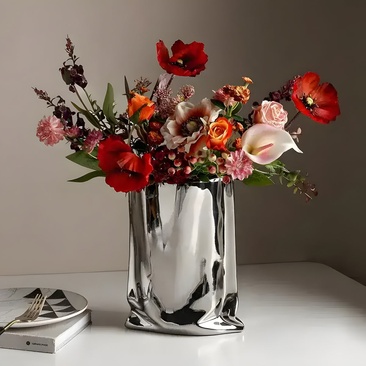Silver shopping bag-shaped vase with red orange pink mixed florals, patterned plate beside
