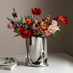 Silver shopping bag-shaped vase with red orange pink mixed florals, patterned plate beside