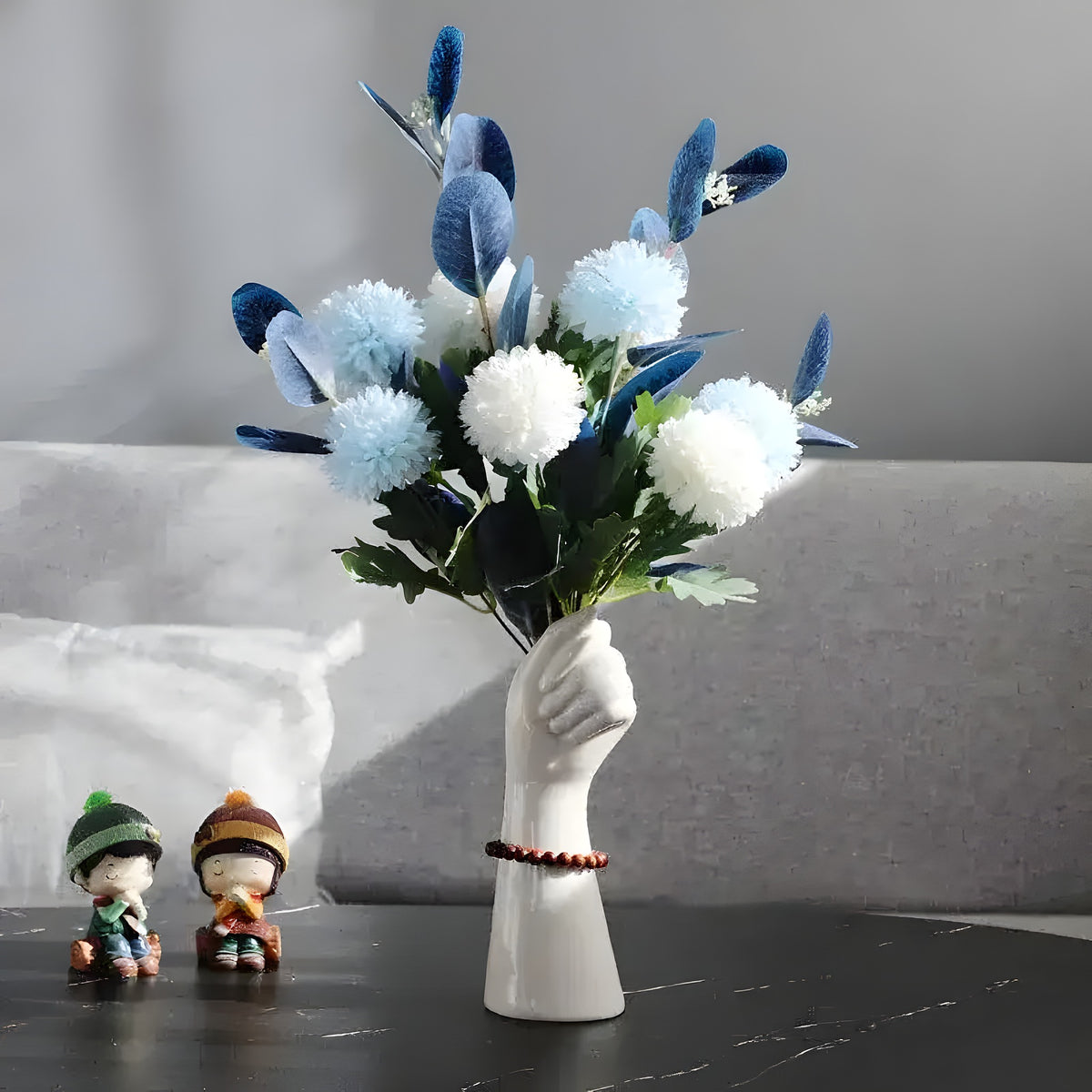 White ceramic hand-shaped vase with bracelet holding blue white pom-pom flowers, marble table