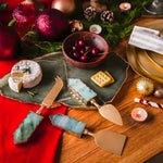 Three green aventurine cheese knives with gold handles styled on slate board with camembert and holiday ornaments