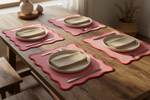 Four scalloped pink table placemats with red edges are arranged on a rustic wooden dining table, each styled with a white plate, beige napkin, silver fork, and silver knife. The scene is lit by warm natural light from a nearby window, with a wooden chair and potted dried plants in the background.