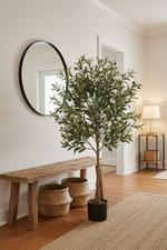 An artificial olive tree in a black pot placed on a wooden floor in a styled entryway, next to a rustic wooden bench, two woven baskets, a round wall mirror, and a side table with a lamp.
