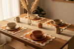 A styled dining table set with beige ceramic bowls, plates, mugs, and cutlery on matching scalloped placemats with orange centers, arranged with a cream runner, glass vase with dried grasses, and a small wooden lidded container in a warm indoor setting.