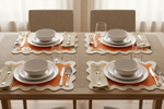 Four place settings arranged on a woven dining table runner, featuring white ceramic bowls and plates, clear drinking glasses, silver cutlery, and orange and beige scalloped placemats. The setting is photographed from above on a light wood table with neutral chairs and soft window light in the background.