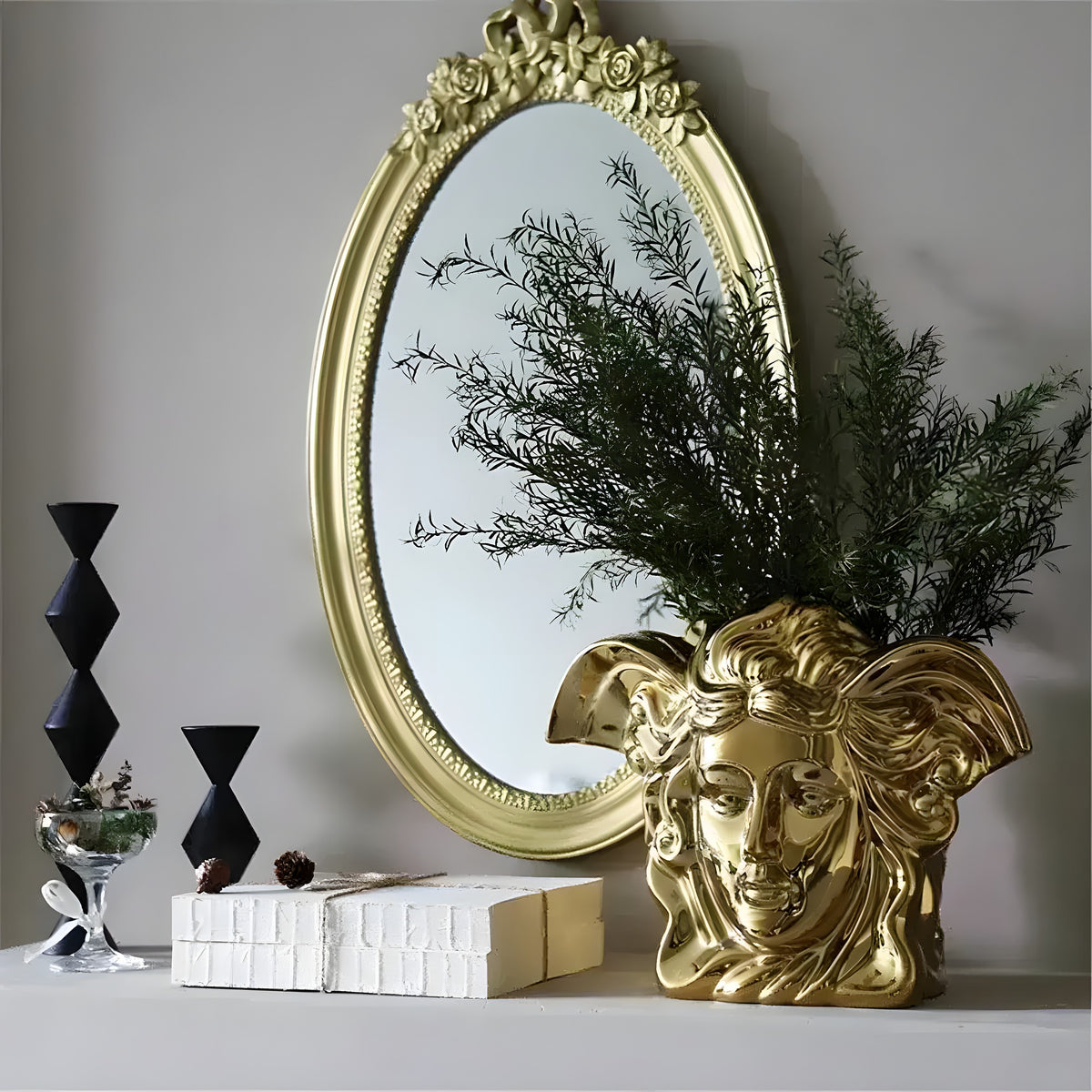 Shiny gold angel face vase holding green branches, styled beside ornate oval mirror and black geometric candlesticks