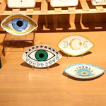 Four eye-shaped porcelain jewelry trays on wood surface: blue, green, moon phase, and sunburst designs with gold trim