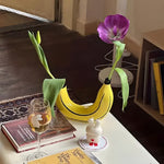 Yellow banana-shaped vase holding two tulip stems on a desk with books and wine glass, in-room setting