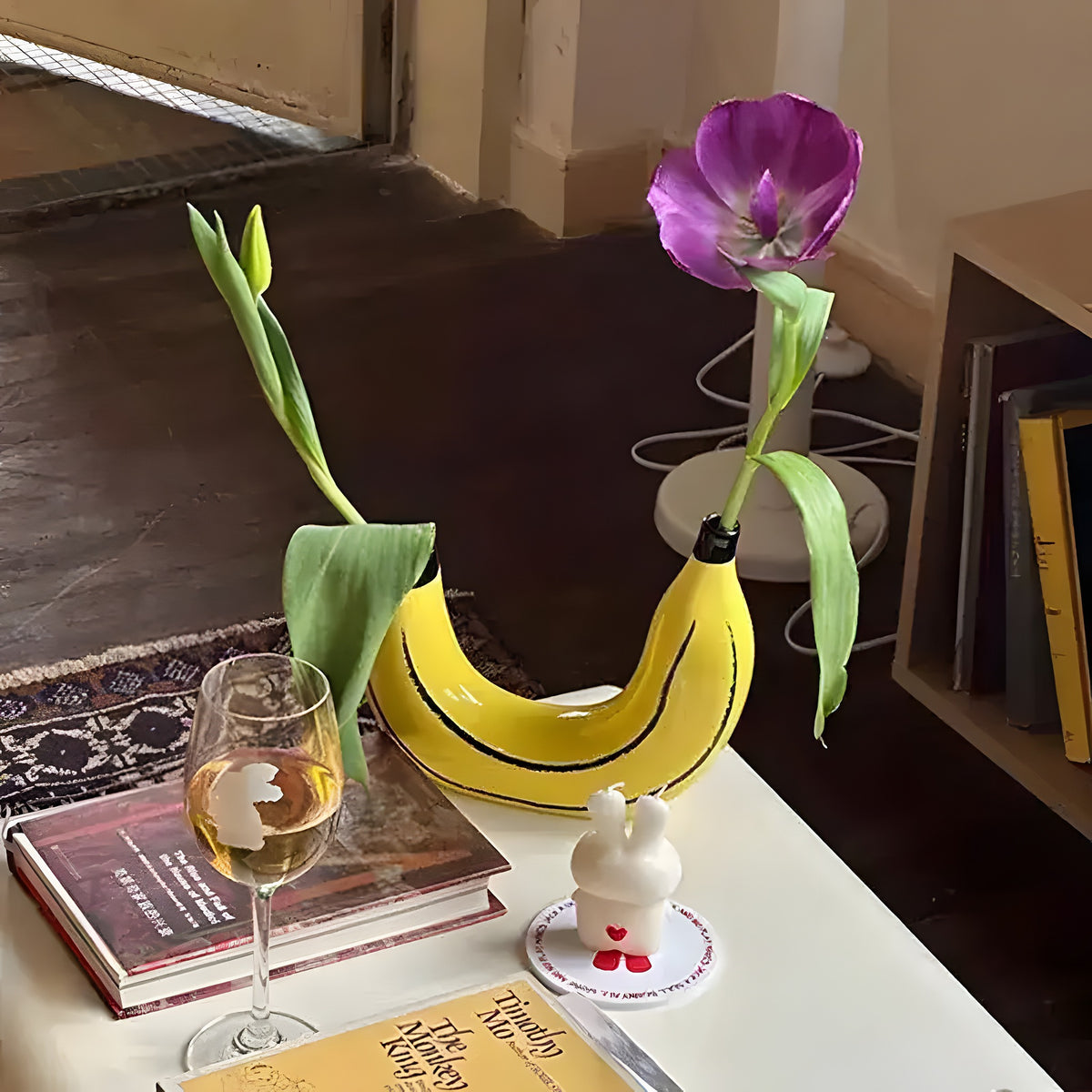 Yellow banana-shaped vase holding two tulip stems on a desk with books and wine glass, in-room setting