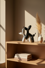 A black and white balloon dog sculpture sits on a light wood shelf next to a small vase with dried pampas grass, with warm sunlight casting shadows across the scene.