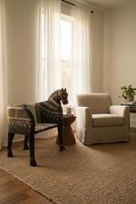 A styled living room corner featuring a beige slipcovered armchair, a decorative horse-shaped bench with a knit patterned throw, a round wooden side table with a mug, and a large textured jute rug on a wood floor.
