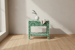 A green and white geometric patterned side table with two shelves, styled with a white ceramic vase containing eucalyptus and a beige throw blanket, set against a textured cream wall and light wood flooring.