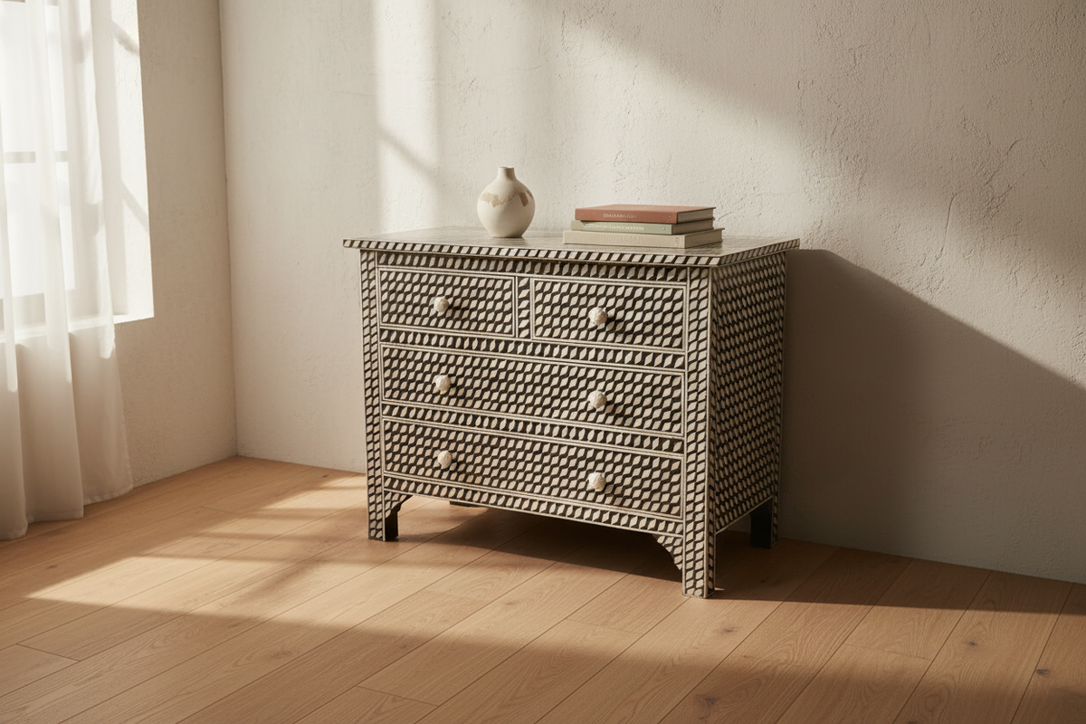 A woven rattan chest of drawers with three drawers and round knobs, styled with a small ceramic vase and books on top, set in a sunlit room with a light wood floor and textured cream walls.